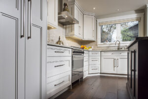 Modern kitchen with white cabinets, stainless steel appliances, stylish kitchen flooring in dark wood, a large window above the sink, and a bowl of fruit on the counter. Natural light brightens the space.