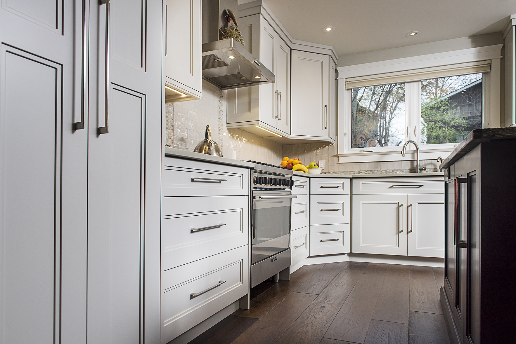 Modern kitchen with white cabinets, stainless steel appliances, stylish kitchen flooring in dark wood, a large window above the sink, and a bowl of fruit on the counter. Natural light brightens the space.