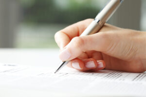 A close-up of a hand holding a silver pen and filling out a home renovation contract on white paper. The background is blurred, drawing focus to the writing hand and the detailed document.