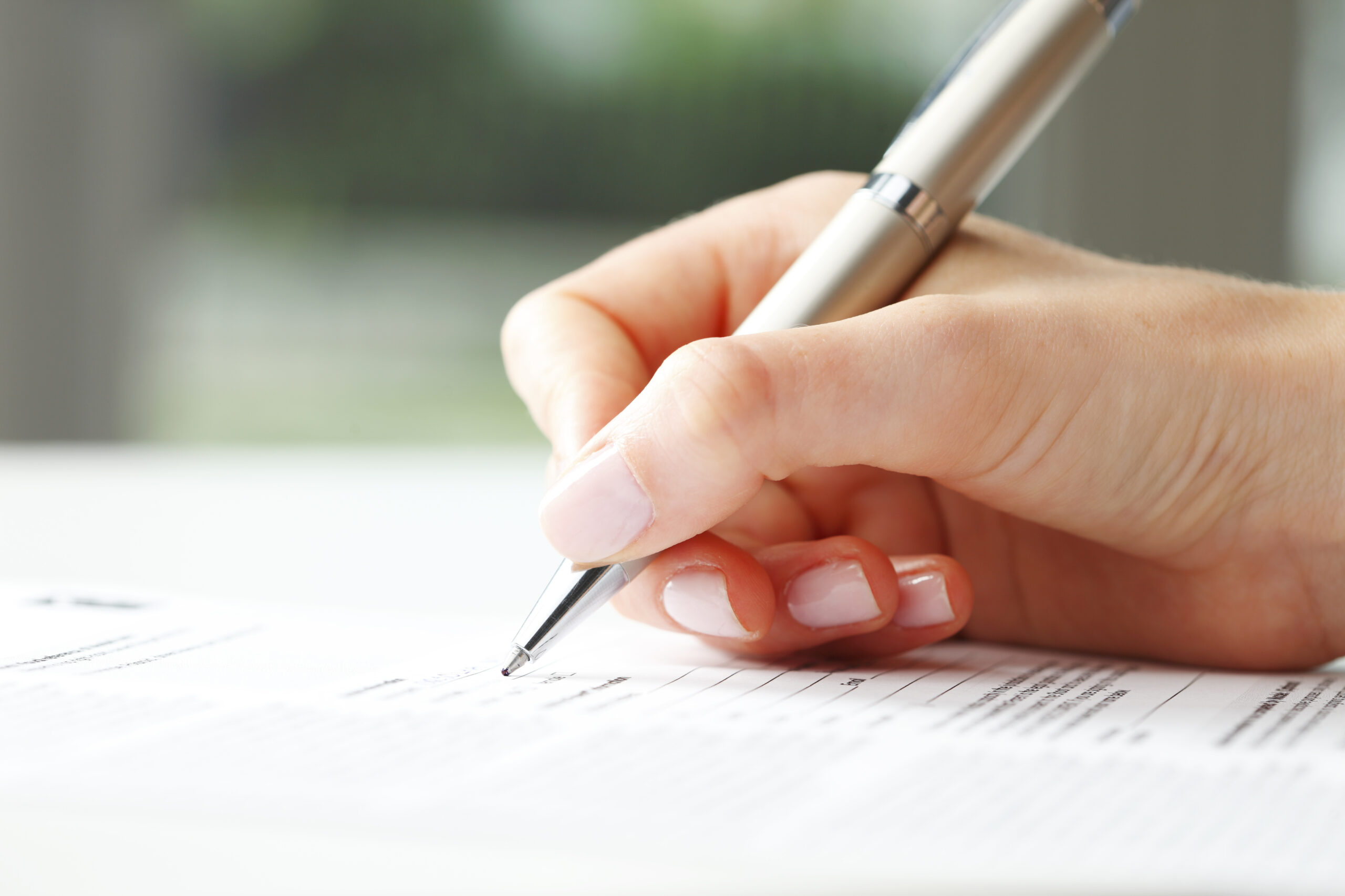 A close-up of a hand holding a silver pen and filling out a home renovation contract on white paper. The background is blurred, drawing focus to the writing hand and the detailed document.