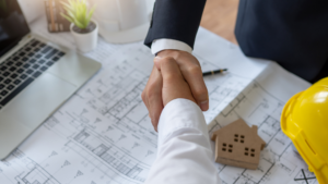 Two people shake hands over architectural blueprints on a table, discussing home renovations, with a yellow hard hat, a small model house, a pen, a plant, and a laptop nearby—raising the important question: should I hire a pro or DIY?.
