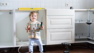 A young child stands in a kitchen under renovation, holding two screwdrivers, with empty cabinets and a power drill on the floor nearby—a playful reminder that even the smallest helpers might appreciate reliable Renovation Warranties when tackling a big reno project.