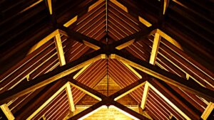 Wooden beams and roof trusses form a geometric pattern on the ceiling of an attic or barn, illuminated by warm yellow light from below—a scene reminiscent of those quiet moments just before the home goes ‘boom’ in the night.