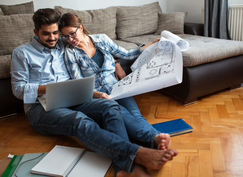 A couple sits on the floor by a sofa, looking at a laptop together. One holds a large blueprint, and notebooks are scattered nearby.