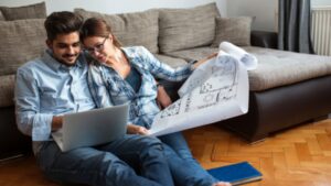 A couple sits on the floor by a couch, looking at a laptop and holding architectural blueprints. They appear to be discussing house plans or renovations in a cozy living room.