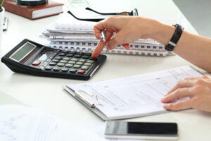 A person presses buttons on a calculator while reviewing documents on a clipboard—perhaps trying to understand a reno quote—with stacked papers, a pen, and a smartphone on the desk.