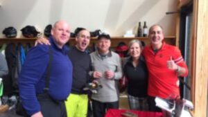 Five adults stand close together indoors, smiling at the camera. They wear casual or sporty clothes, and there are jackets, helmets, and bottles on shelves behind them. One person gives a thumbs up.