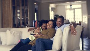 A family of three sits together on a couch, smiling and hugging. The child is snuggled between the two adults, who look happy and relaxed in a cozy living room.