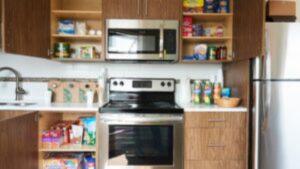 Modern kitchen with brown cabinets, stainless steel appliances, and open shelves displaying boxed and canned food items—much like the inviting spaces at Easter Seals House. Countertops have grocery bags, jars, and a fruit bowl. Refrigerator is on the right.
