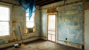 A room with peeling blue paint, exposed ceiling, and a blue tarp hanging down hints at a renovation issue; a broom and mop rest by the wall, an air conditioner sits in the window, while a bucket on the floor catches stray drips. The scene—completed by a screen door leading outside—suggests jaba construction or perhaps even renovation gone wrong.