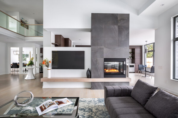 Modern living room with a gray sofa, glass coffee table, fireplace, wall-mounted TV, and open kitchen. Large windows let in natural light, highlighting decor that includes flowers and vases. This inviting space is thoughtfully designed for comfort and style while embracing net zero homebuilding principles for a sustainable future.