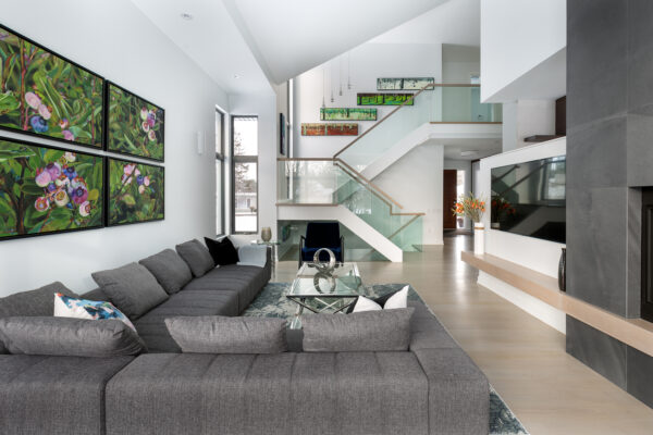 Modern living room with gray sectional sofa, glass coffee table, large wall art, and a sleek fireplace. Open staircase with glass railing and natural light from tall windows highlights the innovative design of this net zero home. Bright, airy space with minimalist decor showcases sustainability at its finest—a true contender for national awards for housing excellence.