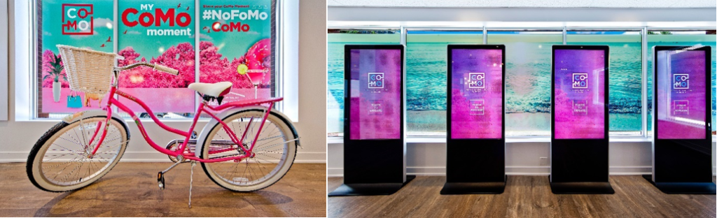 A pink bike with a basket stands indoors in front of colorful “CoMo” posters, capturing the spirit of housing excellence celebrated by the National Awards for Housing Excellence. Next to it, four digital display screens are aligned in front of large windows, showcasing modern home trends.