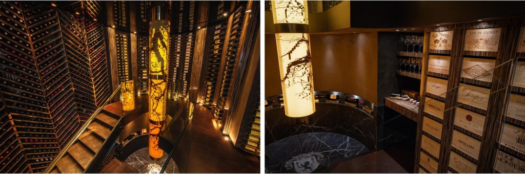 Dimly lit Personalized Wine Cellars feature tall racks of wine bottles, a spiral staircase, and a central column with illuminated artwork—a nod to the latest reno trends in Canada. The tasting area is thoughtfully arranged with wooden wine crates and glasses displayed on shelves, blending style and function for an inviting ambiance.