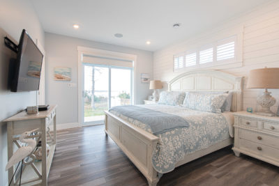 Bright bedroom in a modular home featuring white furniture, light wood floors, a large bed with blue and white bedding, two lamps, a wall-mounted TV, and a sliding glass door opening to an outdoor view. Enjoy modern comfort and style through thoughtful modular home construction.