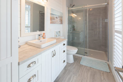 Bright bathroom in a modular home with a white vanity, sink, large mirror, and faucet. A toilet and glass-enclosed shower are in the background. Light wood floor and a blue mat add to the clean, modern look—demonstrating how modular home construction combines style with practicality.