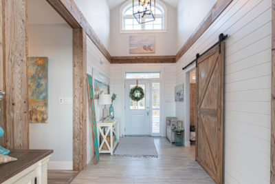 Bright farmhouse entryway with white shiplap walls, wooden accents, a sliding barn door, and a glass front door decorated with a green wreath. Natural light fills the hallway, highlighting rustic and coastal decor—perfect for showcasing the charm and versatility of modular home construction.