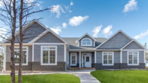 A modern gray modular home with white trim, large windows, and a porch sits under a blue sky. Two leafless trees and a green lawn are in front, showcasing the clean design made possible by modular home construction.