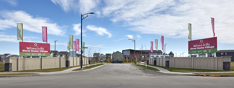 A wide road leads to a neighborhood entrance with colorful flags and large signs reading “Welcome to the Show Home Village.” Reflecting homebuyer preferences highlighted in the CHBA Survey, the inviting scene showcases thoughtfully designed houses lining the background beneath a partly cloudy sky.
