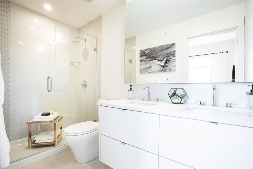 Bright modern bathroom with a glass shower, white double sink vanity, toilet, and wooden stool. Decorative items and a painting add accents to the clean, minimalist design—features that reflect current homebuyer preferences revealed in recent CHBA Survey findings.