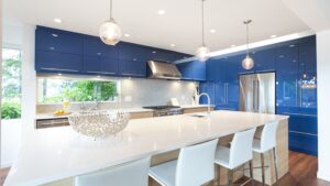 Modern kitchen with glossy blue cabinets, stainless steel appliances, white countertops, a large island with white chairs, pendant lights, and a decorative bowl on the island. Greenery visible through the window creates a welcoming atmosphere—features that consistently rank highly in homebuyer preference studies such as those conducted by Avid Ratings, making this an ideal Kitchen Renovation inspiration.