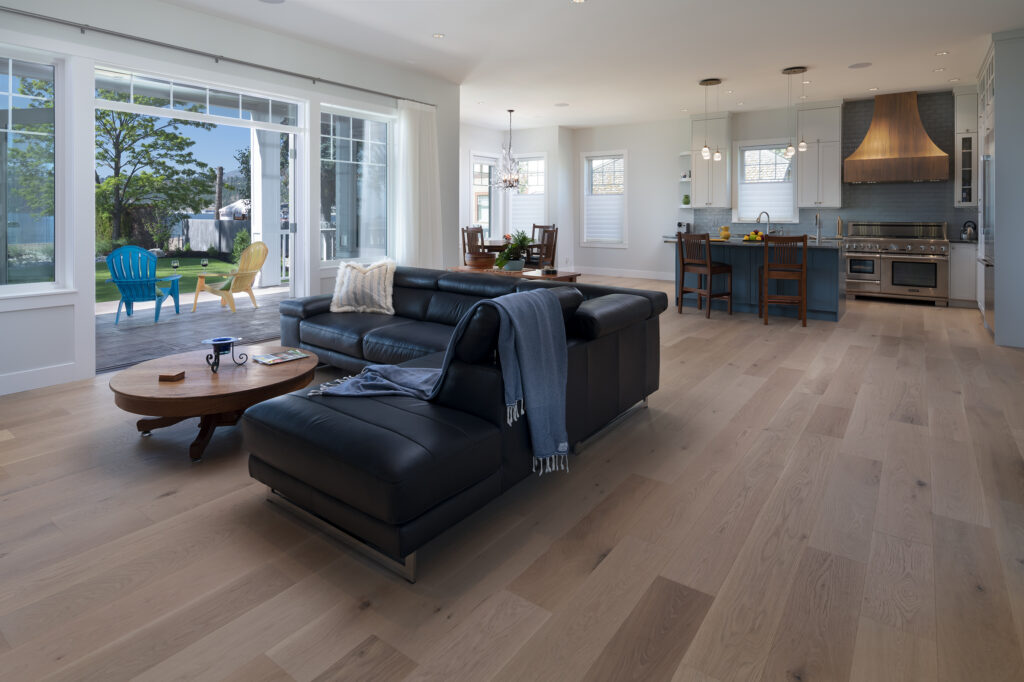 Modern open-concept living room and kitchen with light wood floors, a black sectional sofa, a round coffee table, and large windows showing a patio with Adirondack chairs and a pool in the background—illustrating the comfortable lifestyle possible in net zero ready homes. Discover the difference between net zero & net zero ready homes while enjoying an inviting space filled with natural light.