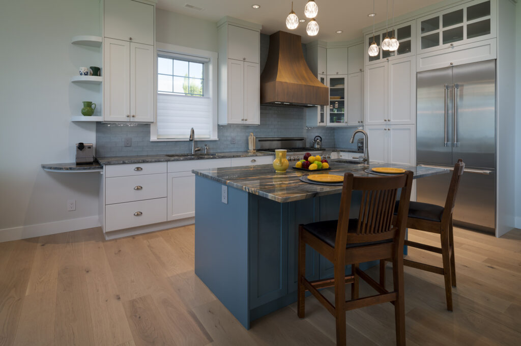 Modern kitchen with white cabinets, a blue island with bar stools, pendant lights, stainless steel appliances, wood flooring, and bowls of fruit and place settings on the island countertop—crafted as part of a net zero ready home that blends contemporary style with energy-efficient design.