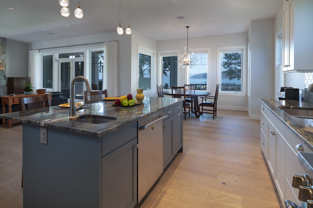 Modern kitchen with a large island, stainless steel appliances, bowl of fruit, and pendant lights; dining area and large windows with a lake view in the background—all within a stylish net zero ready home.
