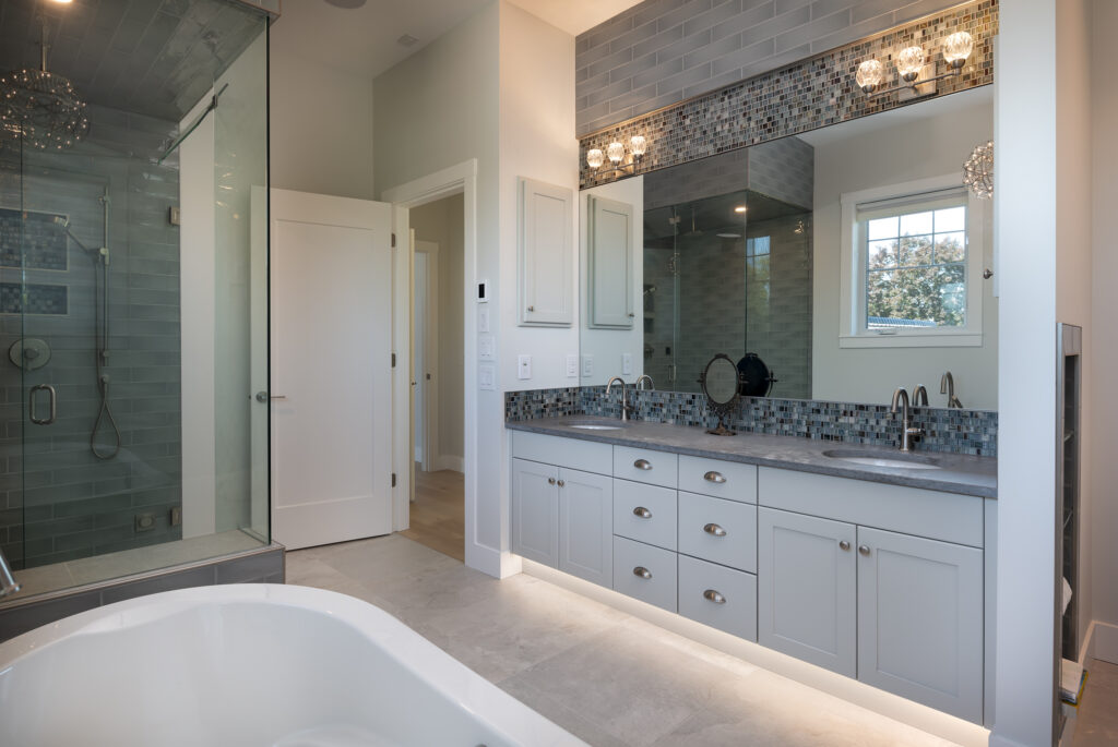 Modern bathroom with a large mirror, double sink vanity, glass shower, white cabinets, gray tile backsplash, bathtub, and a window letting in daylight. Contemporary lighting hangs above the mirror. This elegant space is featured in a net zero ready home—designed for high energy efficiency and sustainability. Discover the difference between net zero & net zero ready homes as you enjoy comfort without compromising on style.