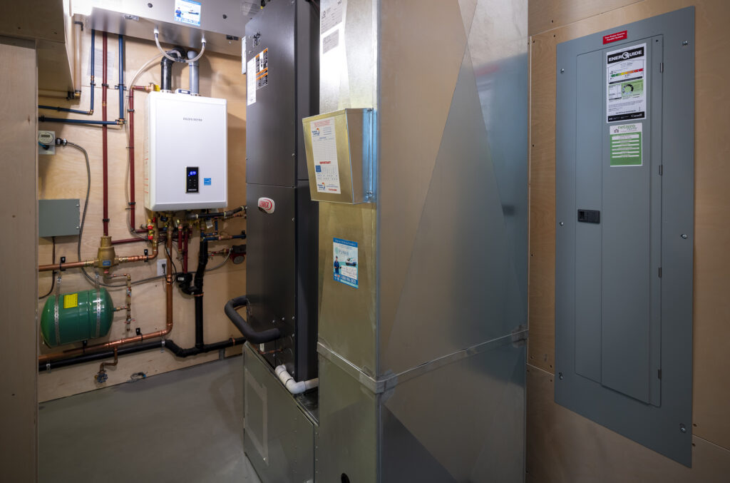 A modern utility room with a furnace, electrical panel, water heater, pipes, and ventilation ducts installed against light-colored walls and floor—designed to support the efficiency of a net zero ready home.