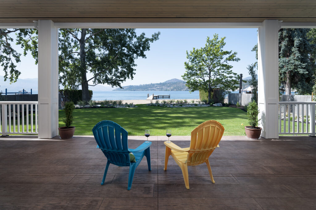 Two colorful Adirondack chairs with glasses of red wine face a lawn, trees, and a lakeside view under a covered patio on a sunny day—a perfect setting to relax and unwind in your net zero home.