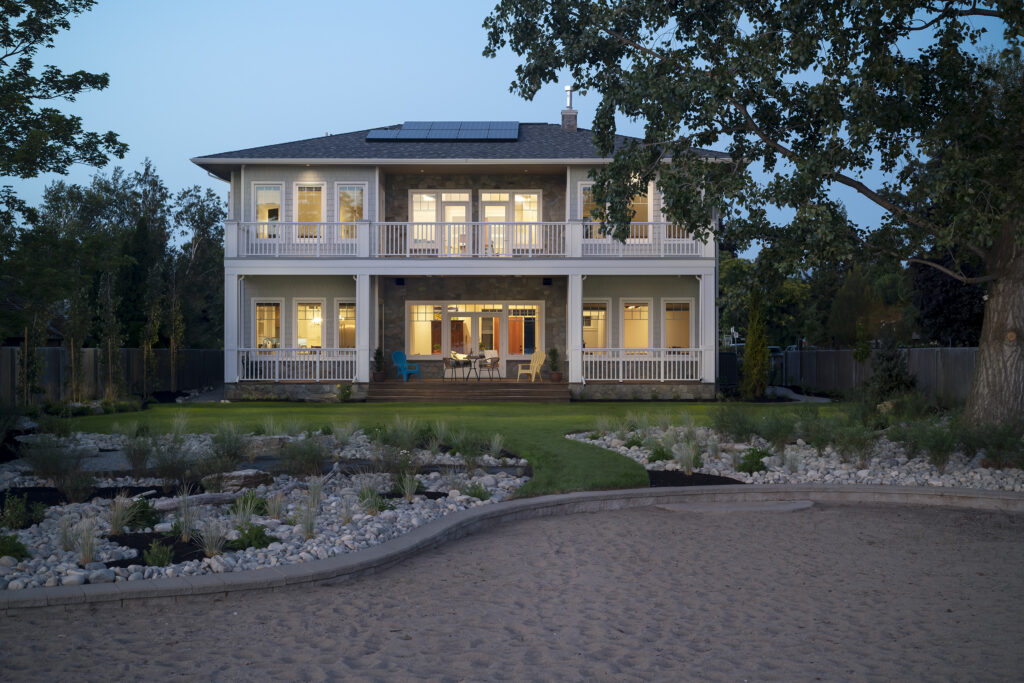 A two-story net zero ready house with stone and siding features double porches with white railings at dusk. The yard includes a manicured lawn, landscaping with rocks and decorative grasses, and a large sandy area in the foreground. Solar panels are on the roof.