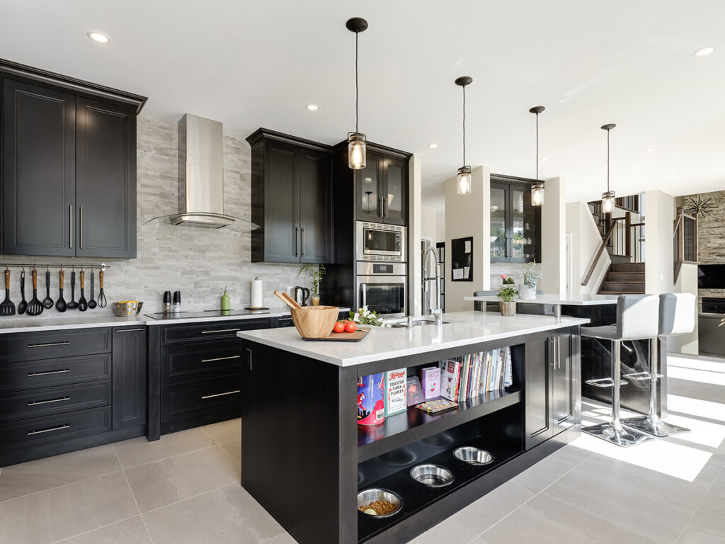 Modern kitchen with black cabinets, stainless steel appliances, and a large island featuring bookshelves, dog bowls, and bar stools. Designed as a net zero ready home, this space showcases pendant lights above the island while sunlight streams through windows.