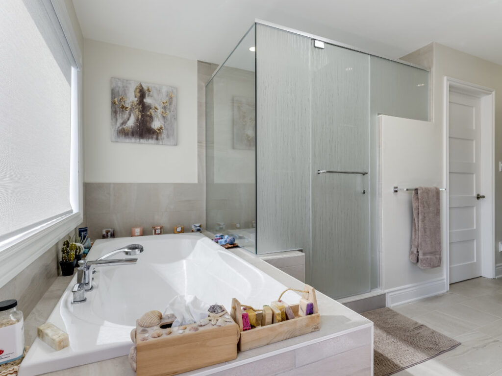 Modern bathroom with a large bathtub, wooden tray holding soaps and accessories, glass-enclosed shower, towels hanging on a wall rack, and artwork above the tub. Natural light brightens the clean, neutral space—perfect for a net zero home designed to maximize comfort while minimizing energy use.