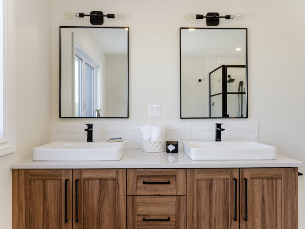 Modern bathroom with a double sink vanity, wood cabinets, two mirrors with black frames, black faucets, tissue box, and folded towels; shower with glass door reflected in mirrors—an elegant space designed for both comfort and style. This home is net zero ready, meaning it’s built to the advanced standards that allow you to easily upgrade to true net zero performance. Discover the difference between net zero and net zero ready homes in this thoughtfully crafted setting.