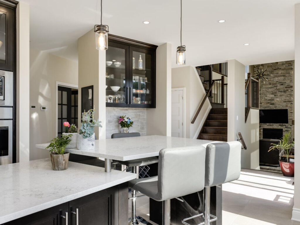 Modern kitchen with white marble countertops, black cabinets, bar stools, pendant lights, and plants. Stairs and a living area with a stone fireplace are visible in the background. Bright natural light fills the space, reflecting the thoughtful design choices of this net zero ready home.