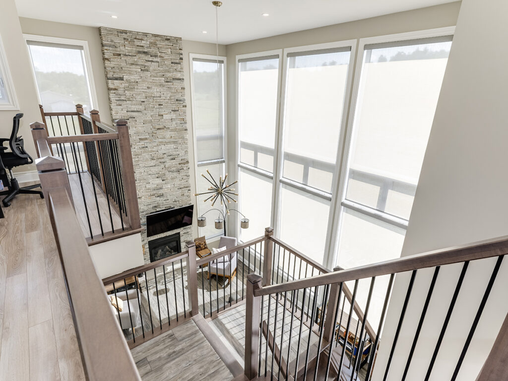 Modern living room with tall windows, a stone fireplace, and a contemporary chandelier. Stairs with wooden railings lead down to a seating area with two chairs and a small table. Bathed in bright natural light, this inviting space reflects the thoughtful design often found in net zero ready homes—built for energy efficiency and future sustainability—highlighting the difference between net zero & net zero ready homes in today’s market.