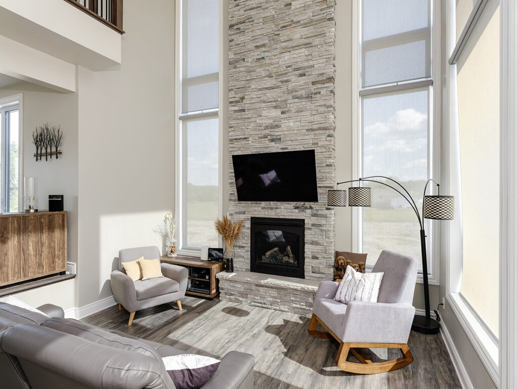 Modern living room with tall stone fireplace, wall-mounted TV, two gray armchairs, gray sofa, floor lamp, large windows, and wood flooring. Minimalist decor with natural light filling the space—a perfect example of comfort and style often found in net zero ready homes. Discover the subtle difference between net zero & net zero ready homes while enjoying energy-efficient living in a space designed for modern lifestyles.