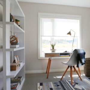 A bright home office with a modern white desk, a white chair, a lamp, and a shelf with books and decor. Sunlight streams in from a large window behind the desk, highlighting the cozy, tidy space—an inspiring spot to learn about sustainable living concepts such as what is net zero ready or the difference between net zero & net zero ready homes.