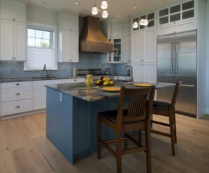 Modern kitchen with blue island, wooden chairs, pendant lights, stainless steel fridge, white cabinets, and a fruit bowl on the countertop. Light wood floors and a window above the sink brighten the space. This inviting design is perfect for those interested in sustainable living and wondering about the Difference Between Net Zero & Net Zero Ready Homes—making it easy to envision an energy-efficient lifestyle right at home.