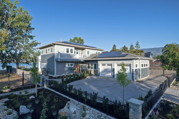 Modern two-story house with solar panels, a large driveway, and a landscaped front yard, surrounded by a black fence, trees, and mountains in the background under a clear blue sky—an ideal example for those wondering what is a net zero home.