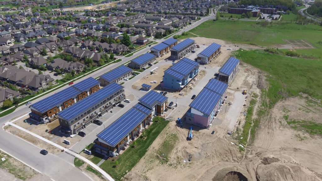 Aerial view of a suburban neighborhood with several new houses, each featuring large solar panels on their roofs, surrounded by grassy fields and ongoing construction. This vibrant community highlights the growing interest in sustainable living, where buyers are increasingly seeking to understand the difference between net zero & net zero ready homes.