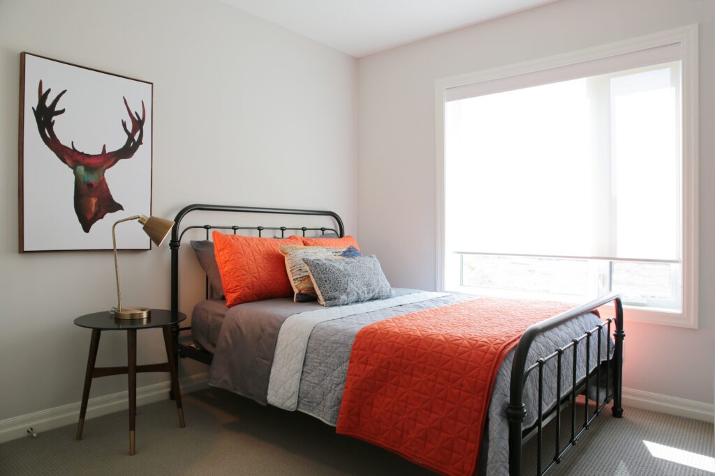 A neatly made bed with orange and gray bedding sits in a bright, net zero ready room, next to a round side table with a lamp and a framed moose art piece on the wall. A large window lets in natural light, highlighting the sustainable features of this energy-efficient space.