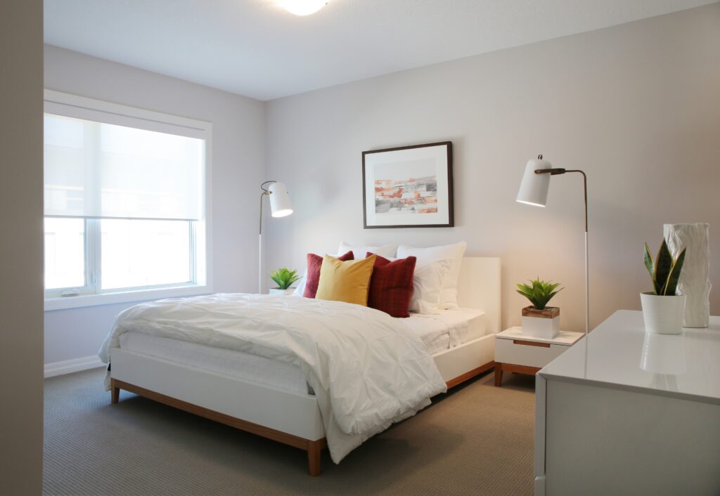 Minimalist bedroom with a modern bed, white bedding, colorful pillows, two bedside tables with plants, tall lamps, framed wall art, dresser, and a large window letting in natural light—perfect for a net zero ready home that prioritizes energy efficiency and sustainable design.