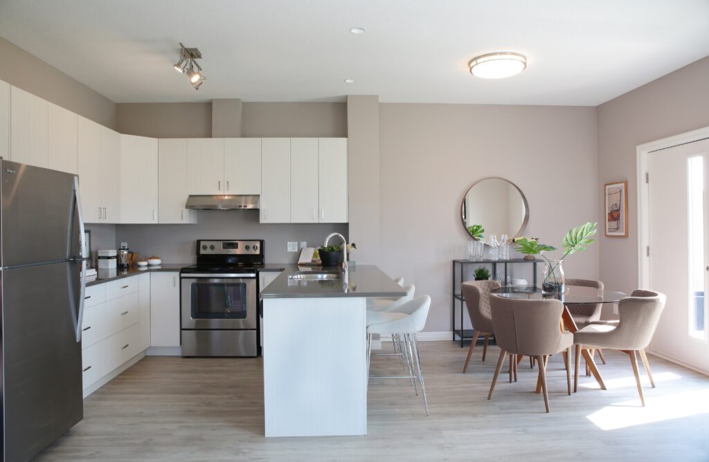 Modern kitchen and dining area with white cabinets, stainless steel appliances, a small round dining table with four chairs, a bar island with stools, and decorative plants. Designed as part of a net zero home, this space features a neutral color palette and natural light, blending energy efficiency with contemporary style.