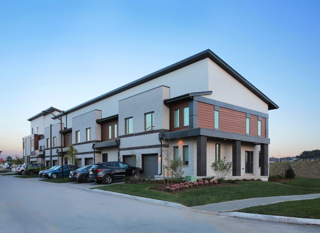 Modern two-story townhouse complex with brick and wood siding, multiple garages, parked cars, landscaped grass, and clear blue sky in the background—these residences are designed as net zero ready homes, blending contemporary style with energy efficiency for sustainable living.