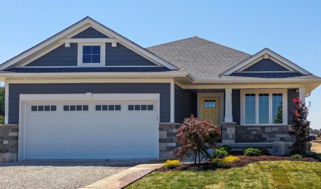 Single-story modern house with gray siding, white trim, a yellow front door, and a two-car garage. The front yard has green grass, shrubs, and a stone pathway under a clear blue sky—an inviting blend of style and sustainability for those seeking Net Zero Homes with innovative design and energy efficiency incentives.