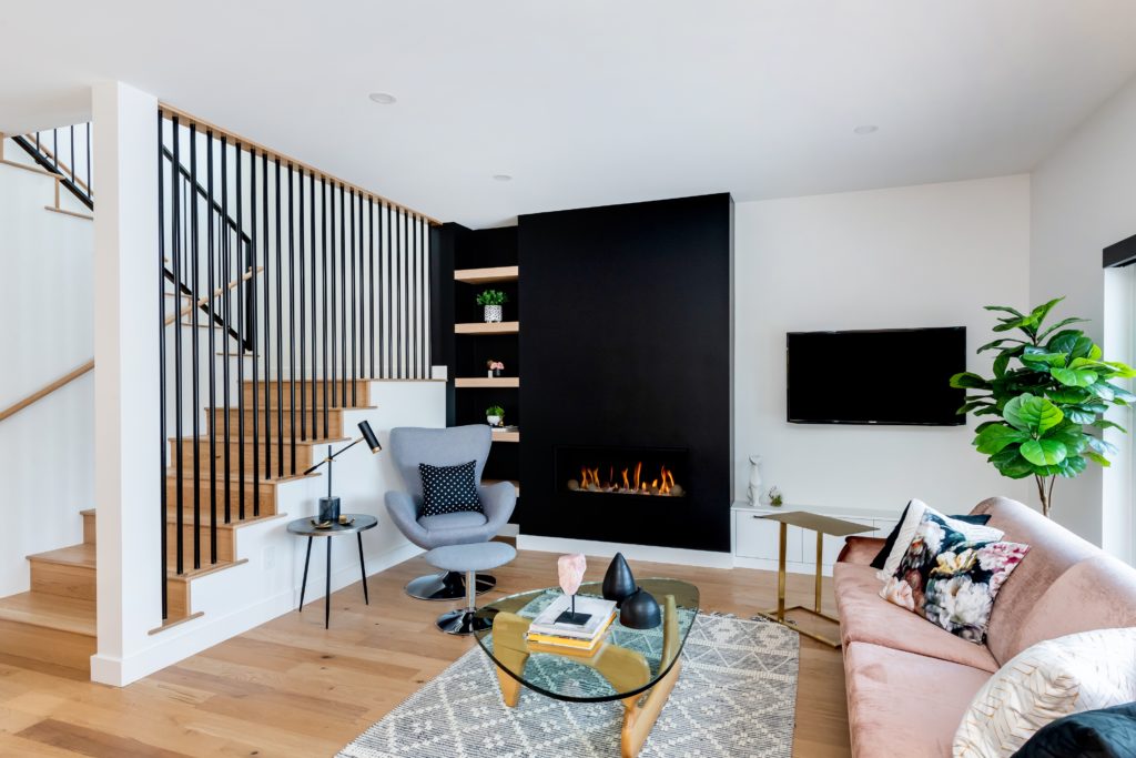 Modern living room with a pink sofa, gray armchair, glass coffee table, fireplace set in a black accent wall, wooden stairs with black railing, TV, and large plant—all thoughtfully designed to balance home wants and essential home needs—on light wood floors and a patterned rug.