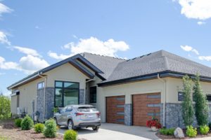 Modern single-story house with stone and beige siding, large windows, and wooden double garage doors. A silver SUV is parked in the driveway on a sunny day with blue skies and scattered clouds. Shrubs line the lawn, creating a welcoming atmosphere that reflects the sustainable design often found in net zero homes.