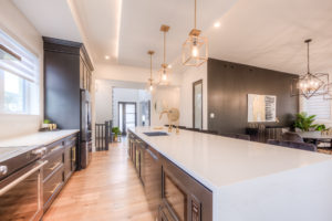 Modern kitchen with a large white island, pendant lights, dark cabinets, wood floor, and an open layout leading to a dining area with black accent walls and decorative plants—designed to complement the energy-efficient lifestyle of net zero homes.